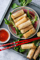Fried spring rolls with red and white sauces, served in china plate on wood tray with fresh green salad and wooden chopsticks over gray blue texture background. Flat lay, close up. Asian food