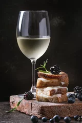  Grilled sandwich with melted goat cheese, blackberry, blueberry, rosemary and honey, served on terracotta board with glass of cold white wine over dark background. Summer appetizer. © Natasha Breen