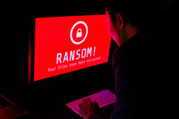 Computer screen with ransomware attack file encrypted alert in red and a man in suit keying on keyboard in a dark room, ideal for online security and digital crime