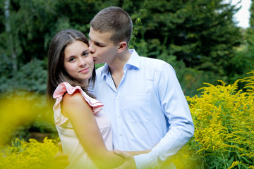 Attractive young couple walking in the garden. Man kissing beautiful brunette womanin cheek. Yellow flowers at hte background