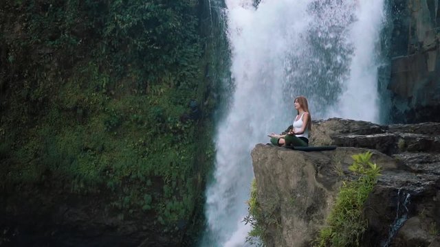Long shot of young slim caucasian woman in white top and green leggings meditating, practicing yoga lotus pose on cliff near Tegenungan Waterfall, Ubud, Bali. Shot with Sony a7s on slider, cloudy day