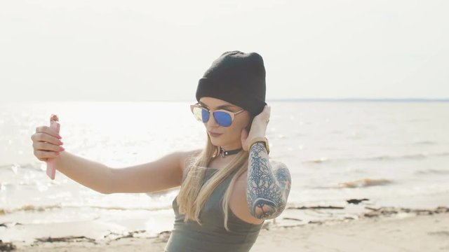 Young blonde with a tattoo in sunglasses and a choker takes off on the phone on the seashore close-up.