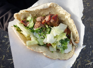 Hand holding traditional greek food, souvlaki, also known as gyros with meat, in pita bread and white paper bag, served at a food festival.