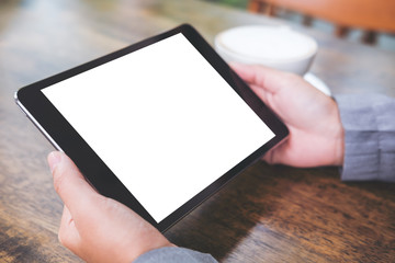 Mock up image of business woman's hands holding black tablet with white blank screen and coffee cup on wooden table in cafe