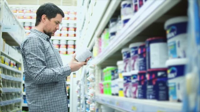 Man in dye shop. Rows of shelves with paint cans. He looking on can and reads label