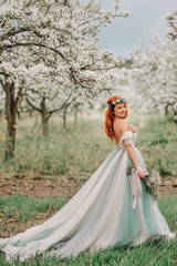 Young red-haired woman in a luxurious dress is standing in a blooming garden