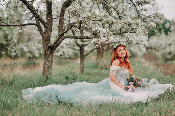 Fototapeta na wymiar Young red-haired woman in a luxurious dress sits on the grass in a blooming garden