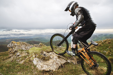 Fototapeta na wymiar Ryder in full protective equipment on the mtb bike climbs on a rock against the backdrop of a mountain range and low clouds