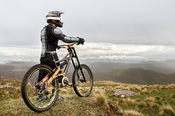 Obraz na płótnie Canvas The rider in the full-face helmet and full protective equipment on the mtb bike stands on a rock against the background of a ridge and low clouds