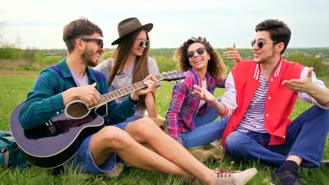 Group of stylish cheerful hipsters with guitar while sitting on a grass in the park. Summer leisure concept. Medium shot