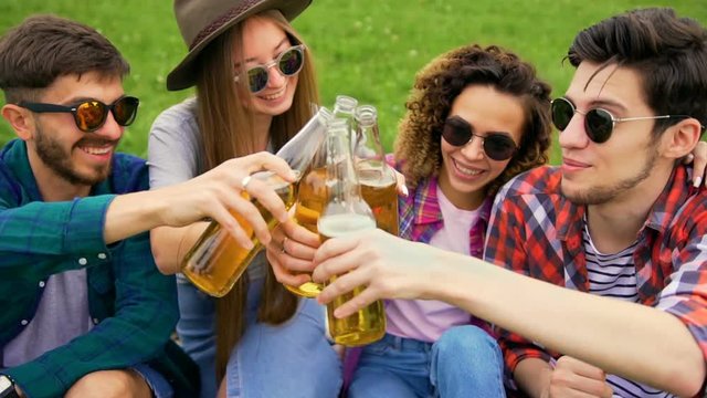 Group of cheerful people are sitting on green grass in the park. They drinks beer and clinking with bottles. Summer lifestyle concept. Medium shot
