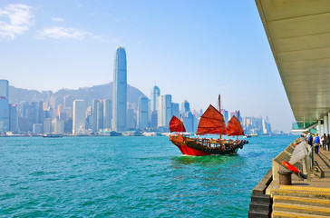 Naklejka premium View of Hong Kong skyline with a red Chinese sailboat passing on the Victoria Harbor in a sunny day.