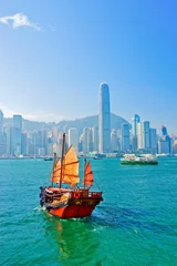 Poster Im Rahmen Blick auf die Skyline von Hongkong mit einem roten chinesischen Segelboot, das an einem sonnigen Tag am Victoria Harbour vorbeifährt. © Javen
