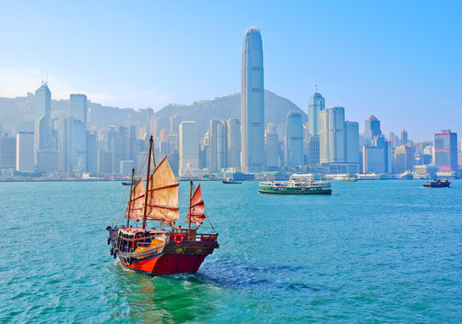 View of Hong Kong skyline with a red Chinese sailboat passing on the Victoria Harbor in a sunny day.