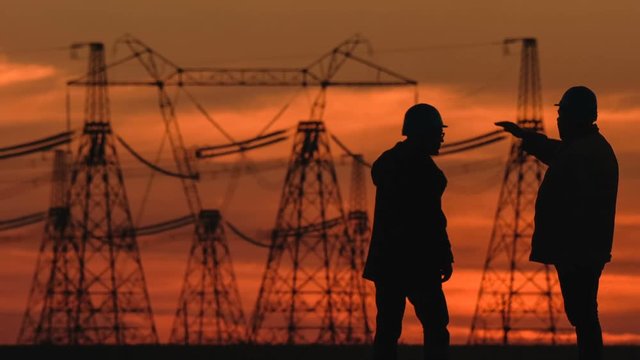 Wide shot of two engineers in hard hat vigorously discussing something at red sunset during an energy substation inspection.