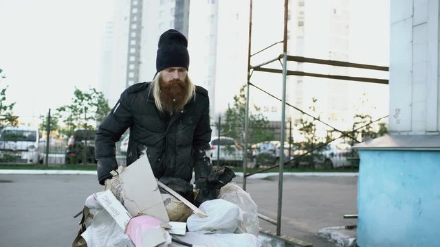 Dolly shot of Bearded homeless man walking down the street with shooping card at cold autumn day