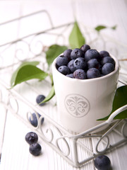 Blueberries and leaves in white ceramic bowl standing in metal basket on white wooden surface