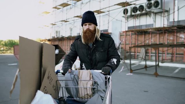 Dolly shot of bearded homeless man walking down the street with shooping card at cold autumn day