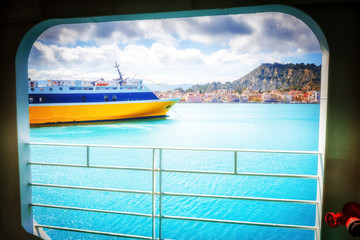 First-person view through port-hole at port of Zakinthos island (Zante traditional name) with yellow-blue ferry moored to jetty.