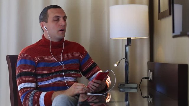Man sits at the table in hotel room and listens to music on his smartphone. Man in sweater with red smartphone with earpieces sits at the table, listens to music and sings a song