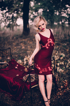 Beautiful and elegant blonde woman with red lips and hair waves wearing wine red nightie posing on the bed outdoors autumn, retro vintage style and fashion.