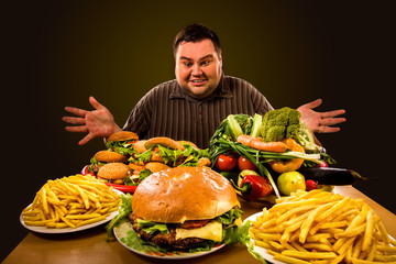 Diet fat man who makes choice between healthy and unhealthy food. Overweight male with hamburgers, french fries and vegetables trays trying to lose weight first time Dinner is field of the working day