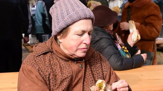 older woman eating a homemade pork  sausage in a bun  with her fingers outdoors, pig slaughter festival, Belo Blato, Vojovina, Serbia, March 12, 2017.
