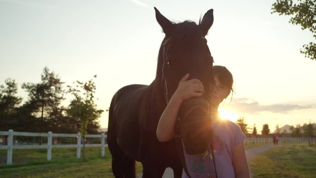 SLOW MOTION, CLOSE UP: Portrait of a cheerful little girl embracing and caressing her dark bay mare at magical golden light sunset. Happy kid hugging and cuddling a colt at the countryside horse ranch
