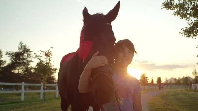 SLOW MOTION, CLOSE UP: Portrait of a cheerful little girl embracing her dark bay mare at magical golden light sunset. Happy kid hugging and cuddling a stunning colt at the countryside horse ranch