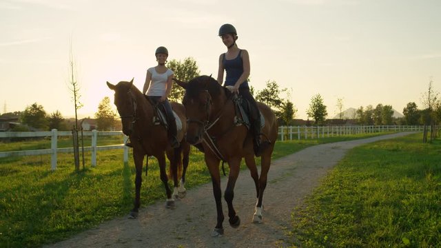 SLOW MOTION, CLOSE UP: Two cheerful young girls horseback riding amazing brown mares walking along the gravel footpath on horse ranch at magical golden light sunset. Friends on relaxing morning ride
