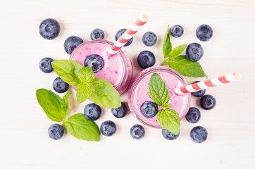 Naklejka na ściany i meble Freshly blended violet blueberry fruit smoothie in glass jars with straw, mint leaves, berries, top view. White wooden board background, copy space.