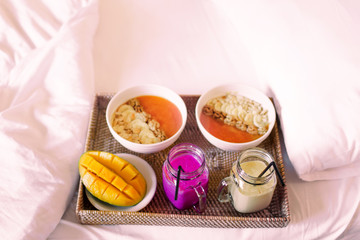 Fresh healthy breakfast in bed for two. Cutted mango, tasty papaya blended smoothie with banana and nuts, tropical fruit shake in jars. All on handmade wooden tray