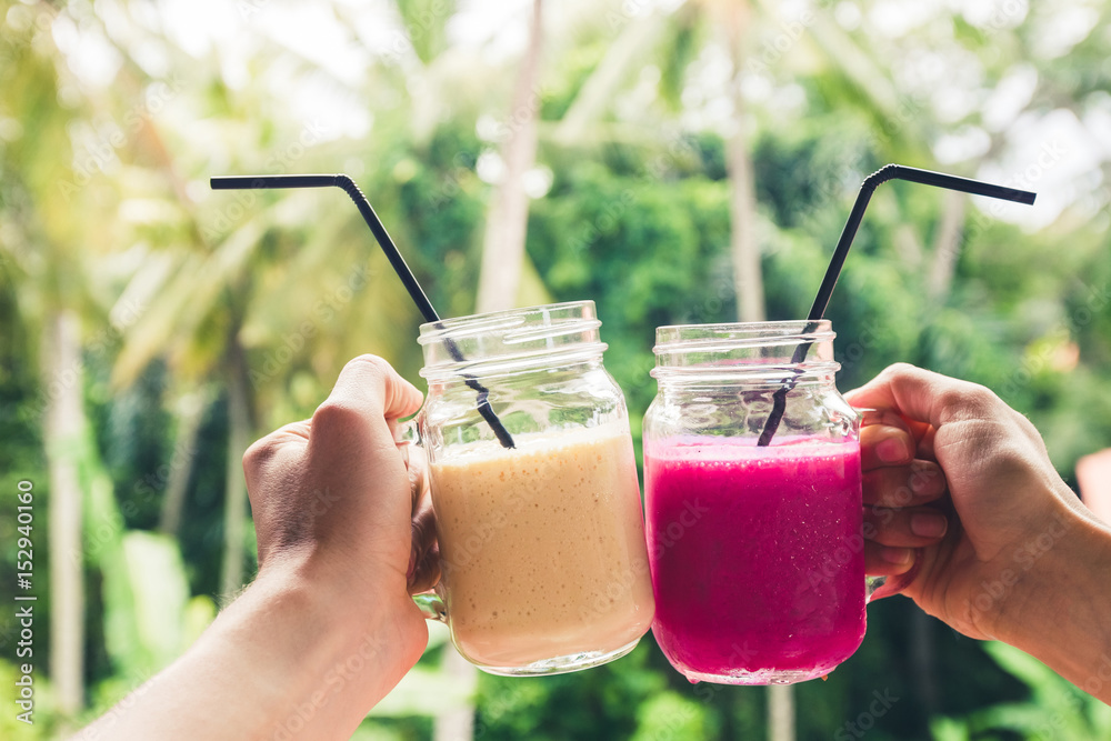 Wall mural Two colorful fruit shakes in hands. Summer and tropical mood. Cold blended drinks, banana and dragon fruit smoothie. Clink glasses by couple hands