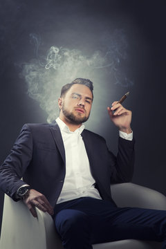 Portrait of bearded young man smoke cigar in modern chair. Wearing suit and shirt