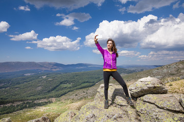 sport hiking or trekking woman with purple jacket, standing on rock peak, with mobile smart phone taking selfie photo picture, behind Lozoya Valley and Guadarrama Park, in Madrid, Spain
