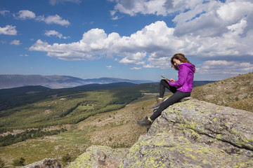 Obraz premium sport hiking or trekking woman with purple jacket, sitting on rock peak, typing in digital tablet, next to Lozoya Valley and Guadarrama Park, in Madrid, Spain 