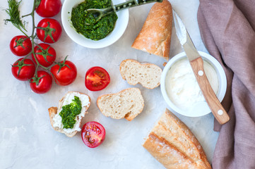 Set of ingredients for bruschettas with goat cheese, pesto sauce and tomatoes abd baguettes on a white stone background. Top view and copy space.