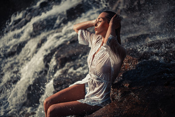 Young woman in white shirt and bikini sits on rock near waterfal