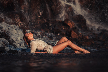 Young woman in white shirt and bikini lies on rock near waterfal