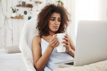 Upset sad beautiful african girl looking at laptop holding cup sitting in chair at home spending her weekend alone.