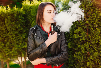 Fototapeta na wymiar Vaping. Young beautiful girl smoking ( vaping ) e-cigarette with smoke outdoors. Vapor concept.