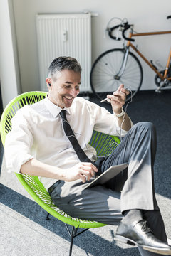 Smiling businessman sitting in an armchair using tablet, smartphone and earphones