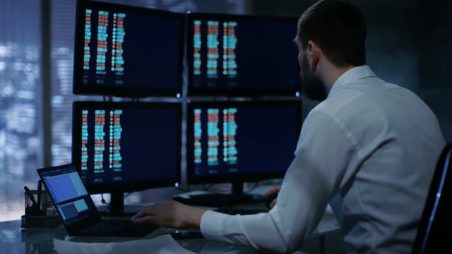 Late at Night Trader Reads Numbers on His Multiple Displays with Stock Information on Them. In Background Big City Window View. Shot on RED EPIC-W 8K Helium Cinema Camera.