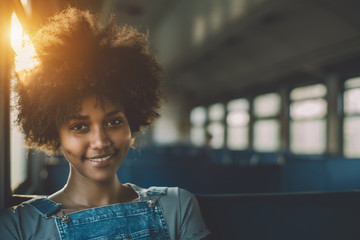 Young smiling beautiful black girl with curly afro hair sitting goes by suburban train, attractive mixed teenage female on seat of empty electrical train with area for your text message or logo - Powered by Adobe