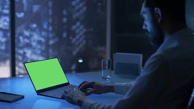  Late at Night Businessman Works on a Laptop with Green Mock-up Screen in His Private Office with Big City Window View.Shot on RED EPIC-W 8K Helium Cinema Camera.