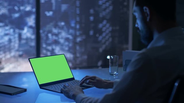 Late at Night Businessman Works on a Laptop with Green Mock-up Screen in His Private Office with Big City Window View. Shot on RED EPIC-W 8K Helium Cinema Camera.