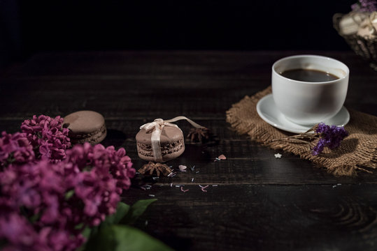 Cup of coffee on linen napkin and several macarons in braid vase with branch of lilac