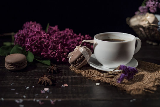 Cup of coffee on linen napkin and several macarons in braid vase with branch of lilac