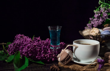 Cup of coffee on linen napkin and several macarons in braid vase with branch of lilac and pile of absinthe