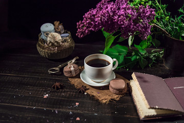 White cup with coffee on a linen napkin and old wooden table and stack of macarons. Old book and lilac.